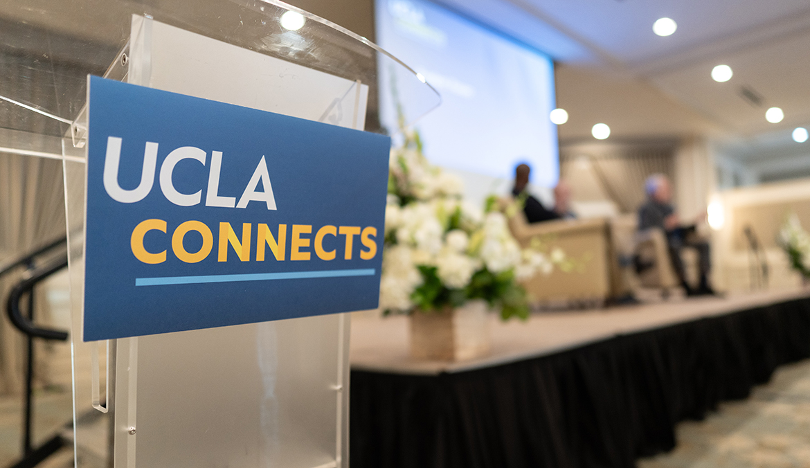 Alt text / caption: A clear acrylic podium in the foreground displays a blue sign reading "UCLA Connects" in white and gold text. 