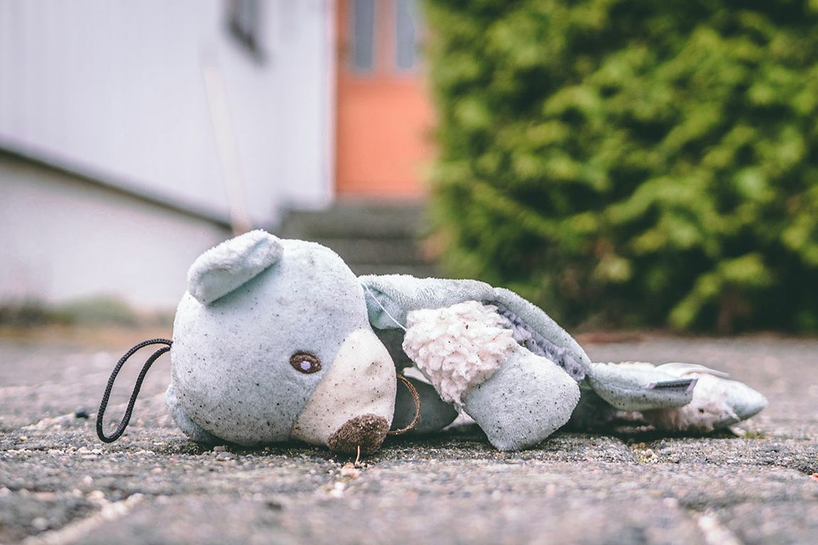 A worn and weathered stuffed animal toy lies abandoned on a concrete surface outdoors, with a blurred building and greenery in the background.