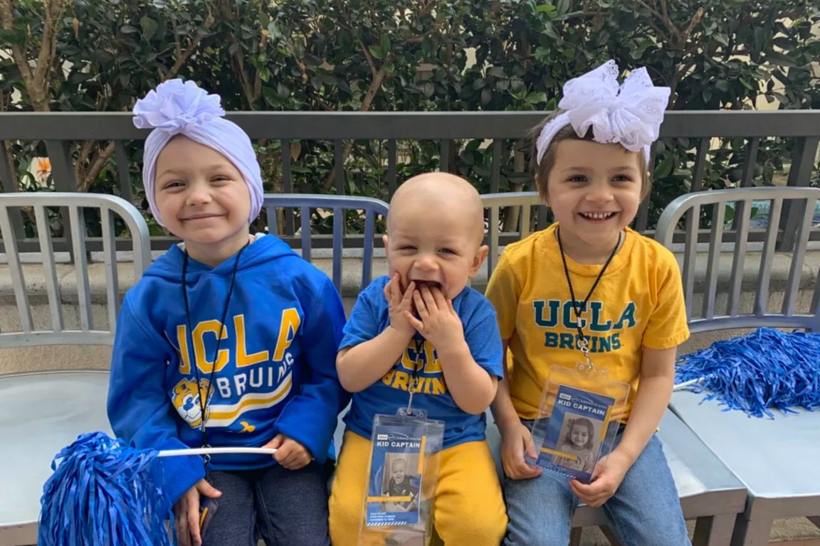Three young children wearing UCLA Bruins gear and "Kid Captain" lanyards smile while seated together at a UCLA Health event, with blue pom-poms visible nearby.