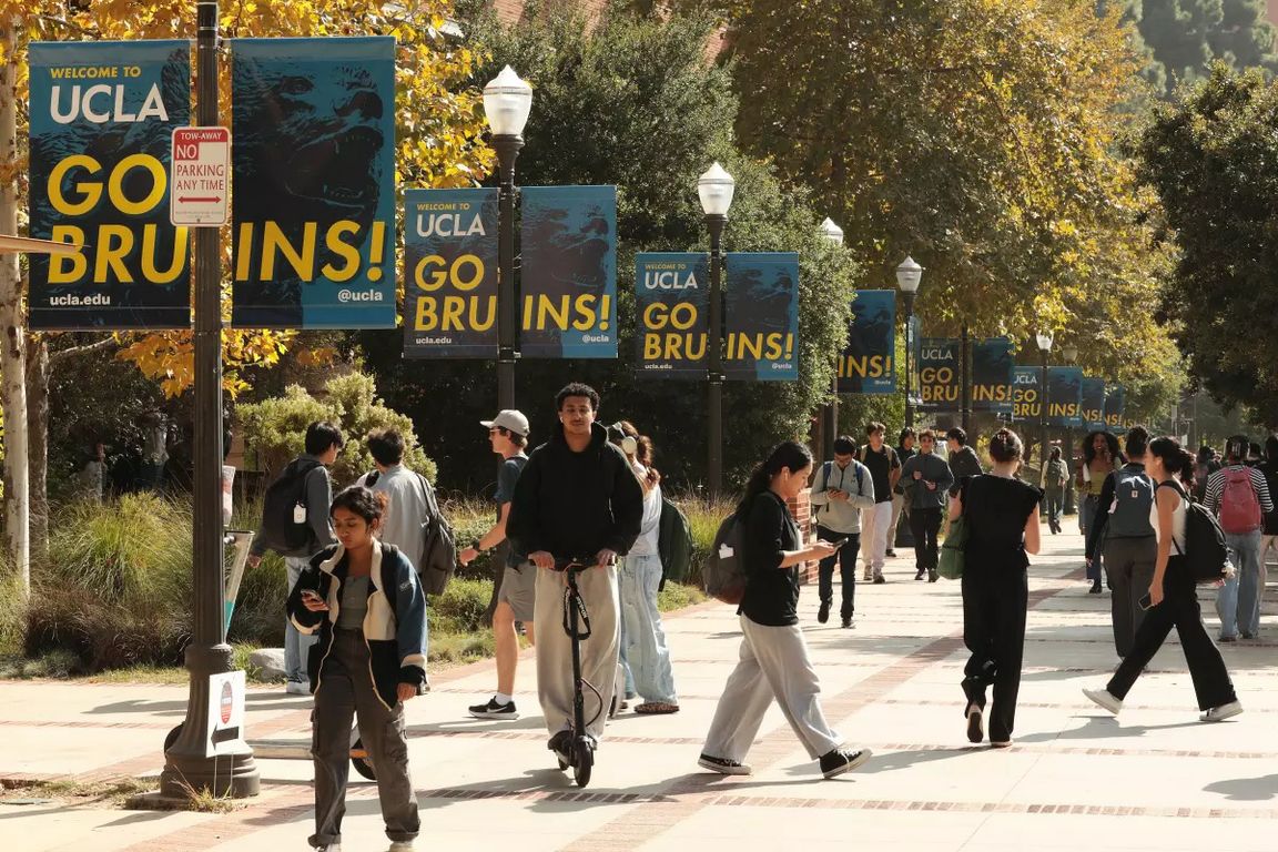 Students walk and scooter along a tree-lined campus pathway at UCLA, flanked by blue and gold "Welcome to UCLA — Go Bruins!" banners. Los Angeles Times owned photo.