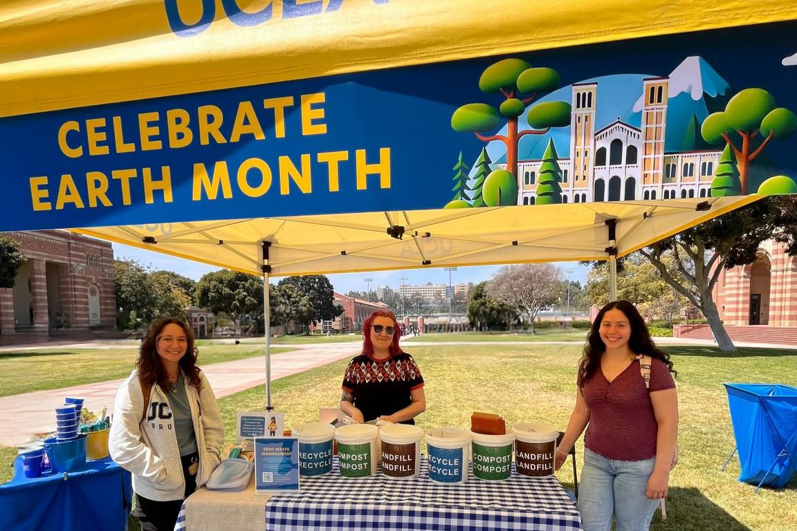Three people smile at a UCLA Earth Month outdoor tabling event, with recycling, compost and landfill sorting bins displayed on the table beneath a "Celebrate Earth Month" banner.