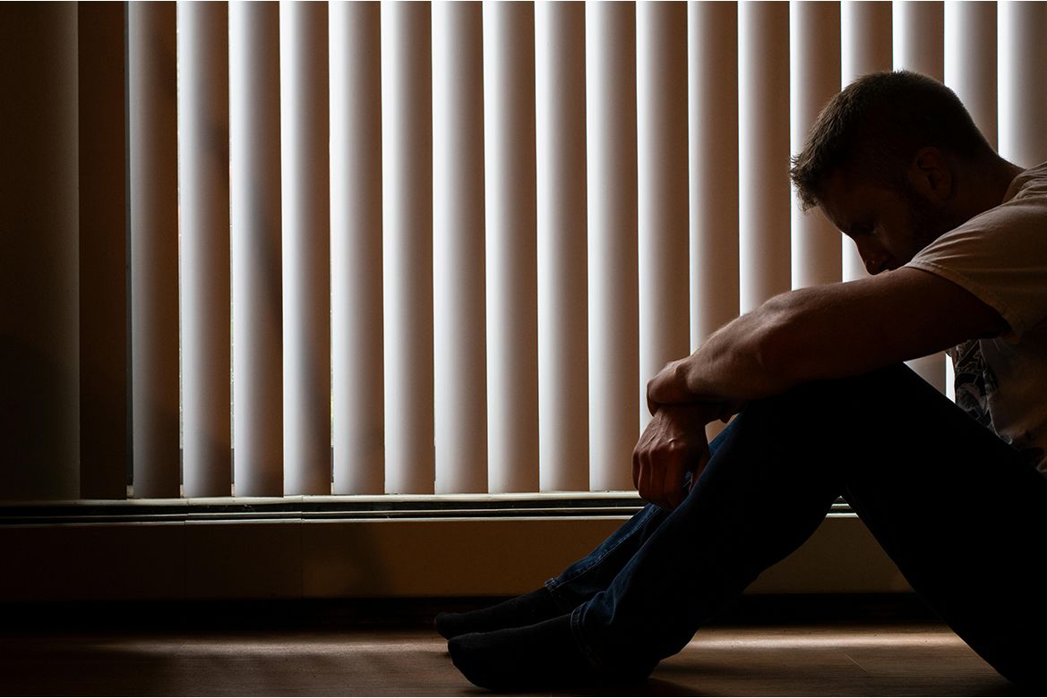 Man sitting in front of closed blinds looking down and hugging knees