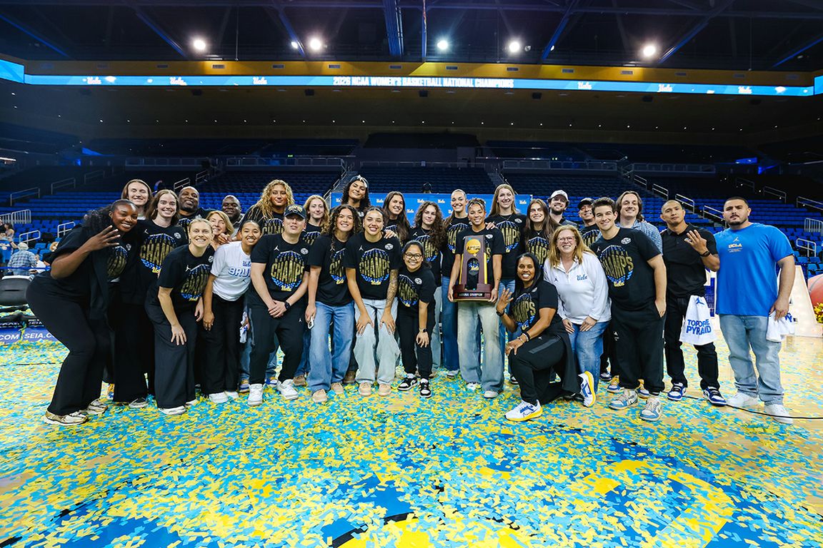 The UCLA women's basketball team, coaches, and staff pose with the NCAA Championship trophy on a confetti-covered Pauley Pavilion court, celebrating their 2026 National Championship title.