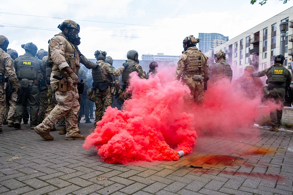 Federal officers wearing gas masks standing near red smoke billowing from a tear gas canister