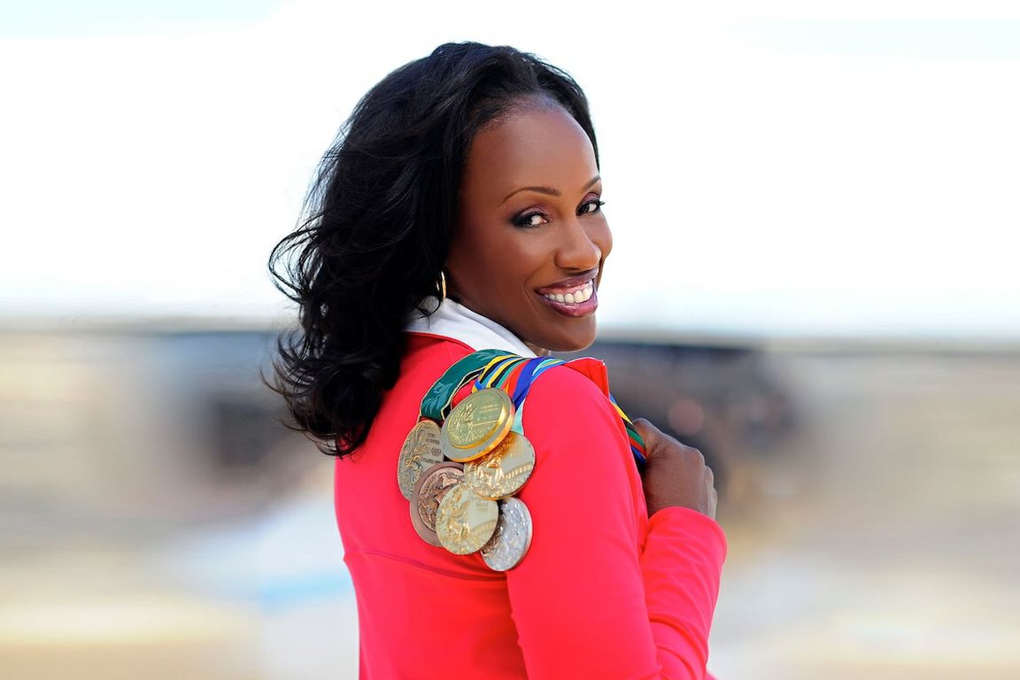 Jackie Joyner-Kersee in red jacket, looking back over her shoulder while holding Olympic medals