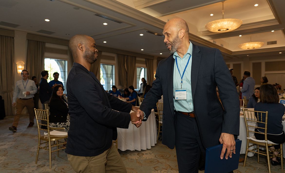 Two men shake hands and smile in a well-lit banquet hall during a professional event. The man on the right wears a navy blazer, light blue dress shirt, and a conference lanyard with a name badge.