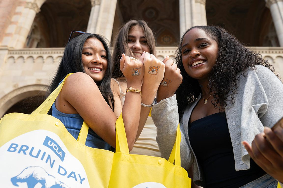 Three smiling students hold up their fists to display matching UCLA Bruin bear temporary tattoos on their hands during UCLA Bruin Day. 