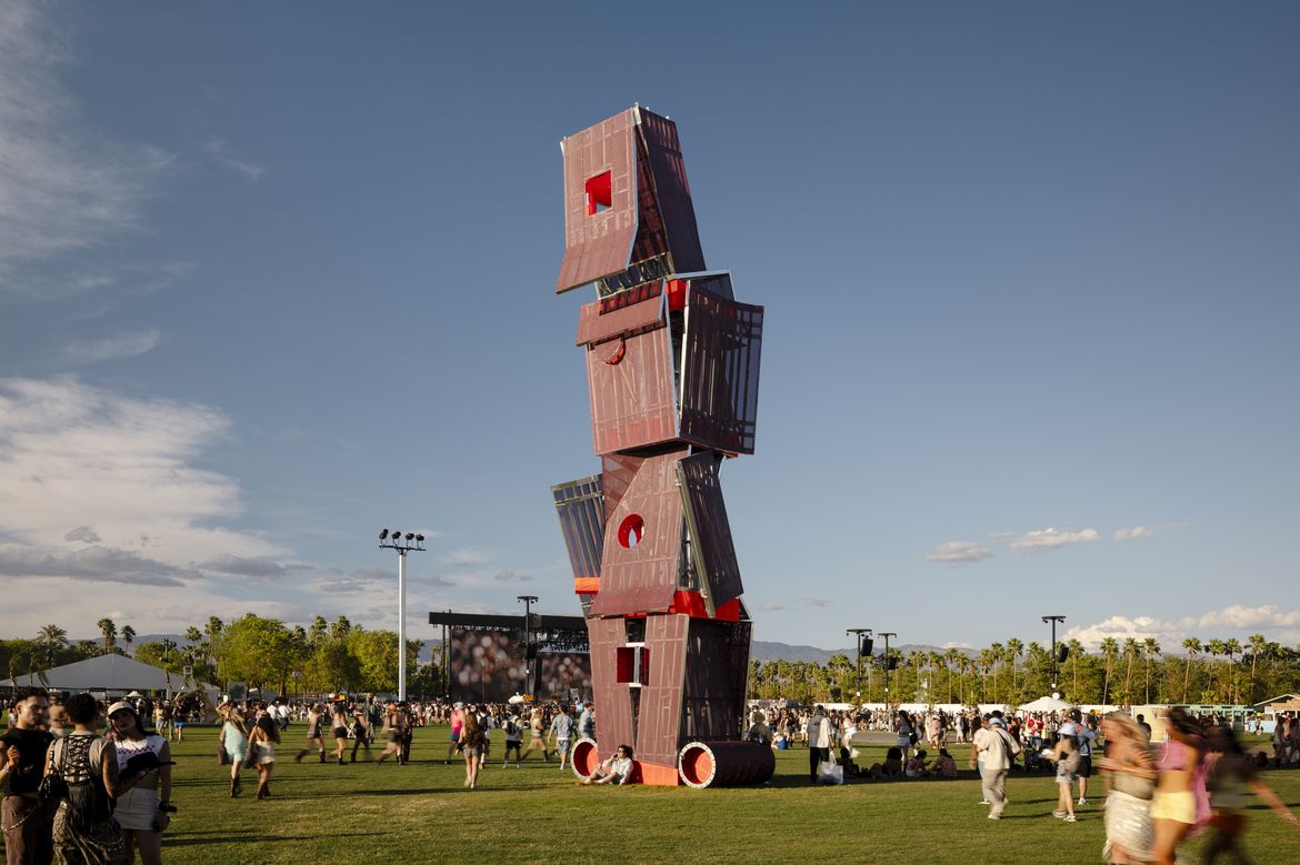 A "tower" made of four geometric boxes in the middle of a field as people walk by