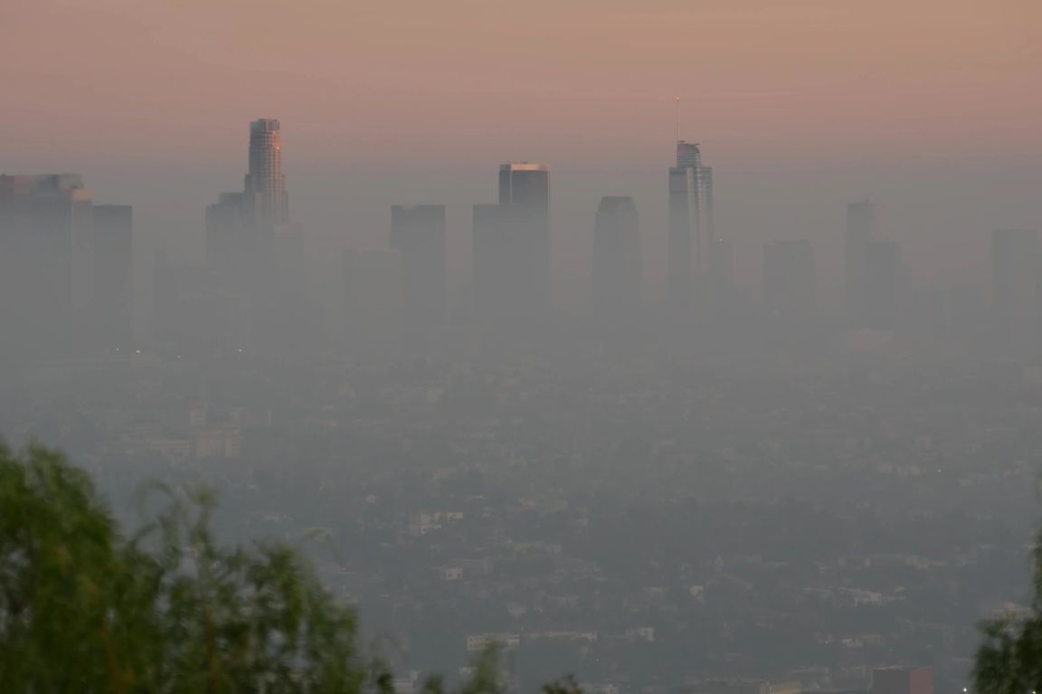 The Los Angeles skyline shrouded in a thick layer of smog at dusk, with skyscrapers barely visible through the haze beneath a pink-tinged sky. For KCRW use only