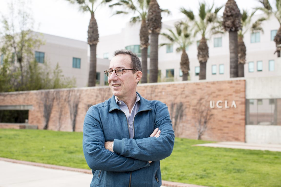 David Eisenman wearing glasses and a blue jacket stands with arms crossed in front of a UCLA brick sign and palm trees on campus.