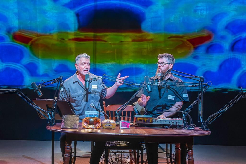Two men wearing blue collared tops sitting at a table with a model train and other artifacts