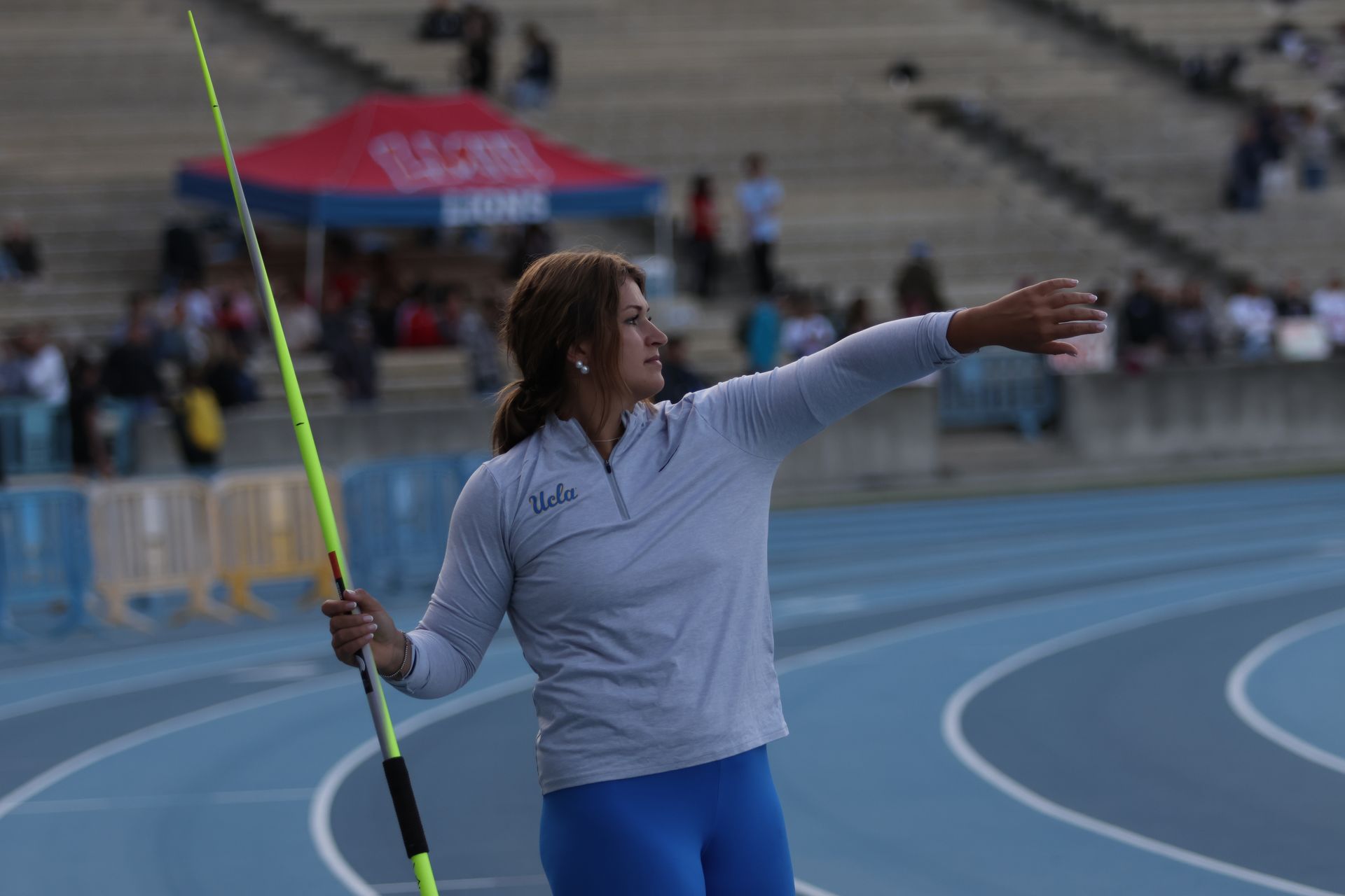 A female athlete in a grey UCLA quarter-zip and blue leggings prepares to throw a neon yellow javelin.