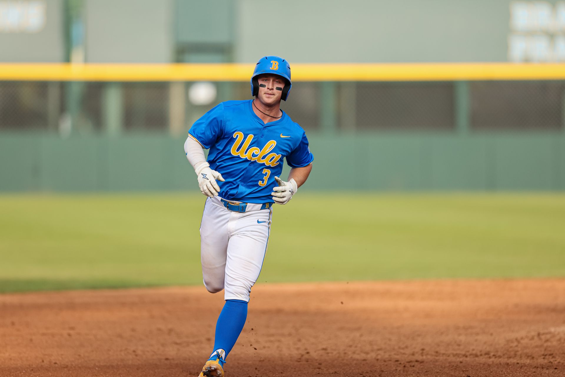 A UCLA baseball player in a blue jersey and helmet runs across the dirt infield during a game.
