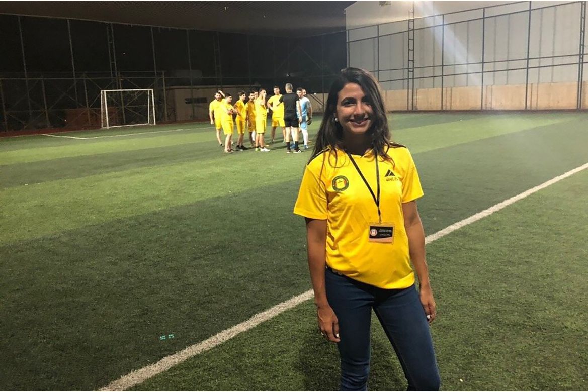 Midrange shot of Salma Mousa, in yellow soccer jersey and dark pants, standing on a soccer field with a group of players in the background 