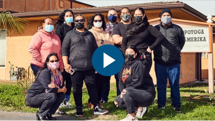 A group of masked community members pose outside a building on a sunny day near a building.