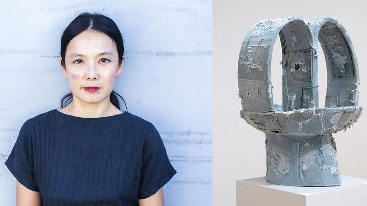 Artist Anna Sew Hoy stands beside one of her sculptural works displayed on a pedestal against a light-colored gallery wall.