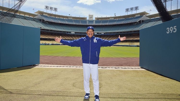 Dave Roberts stands on the field at Dodger Stadium with his arms outstretched, wearing a Los Angeles Dodgers jacket and cap.