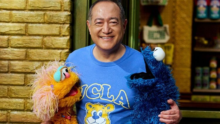 Alan Muraoka smiles while standing between two colorful Sesame Street puppets, wearing a blue UCLA T-shirt.