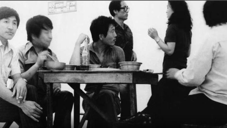 A black-and-white photo shows several people gathered around a small table, seated and standing as they talk in a modest indoor room.