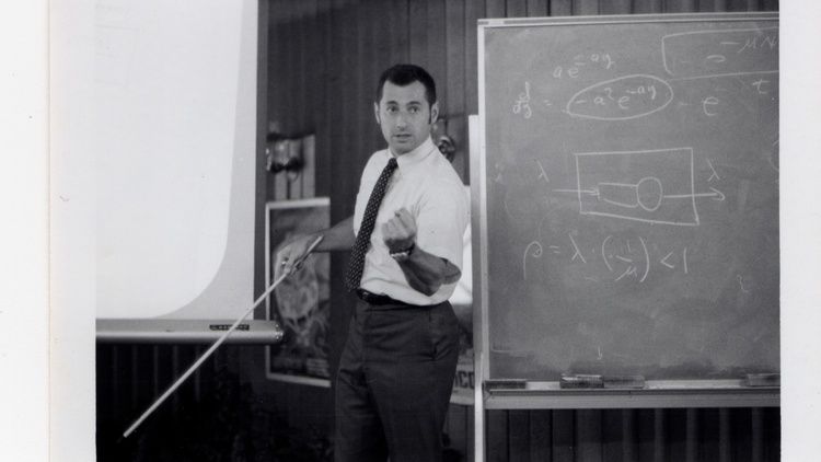 A black-and-white photo shows Leonard Kleinrock in 1970 standing beside a chalkboard, gesturing with a pointer as equations and diagrams are written behind him.