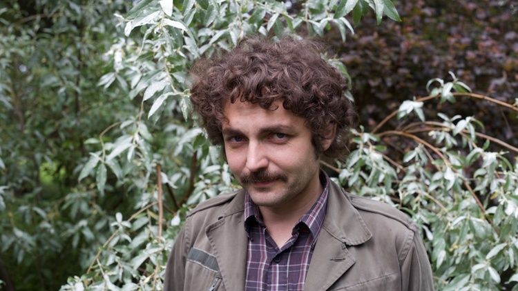 Boris Dralyuk standing in front of green foliage while wearing a green jacket over a plaid shirt.