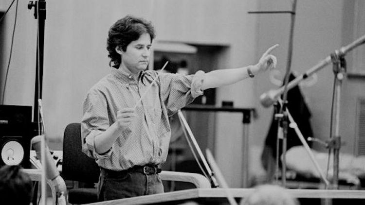 Composer James Horner conducts musicians during a recording session, raising one arm while holding a baton amid microphones and studio equipment.