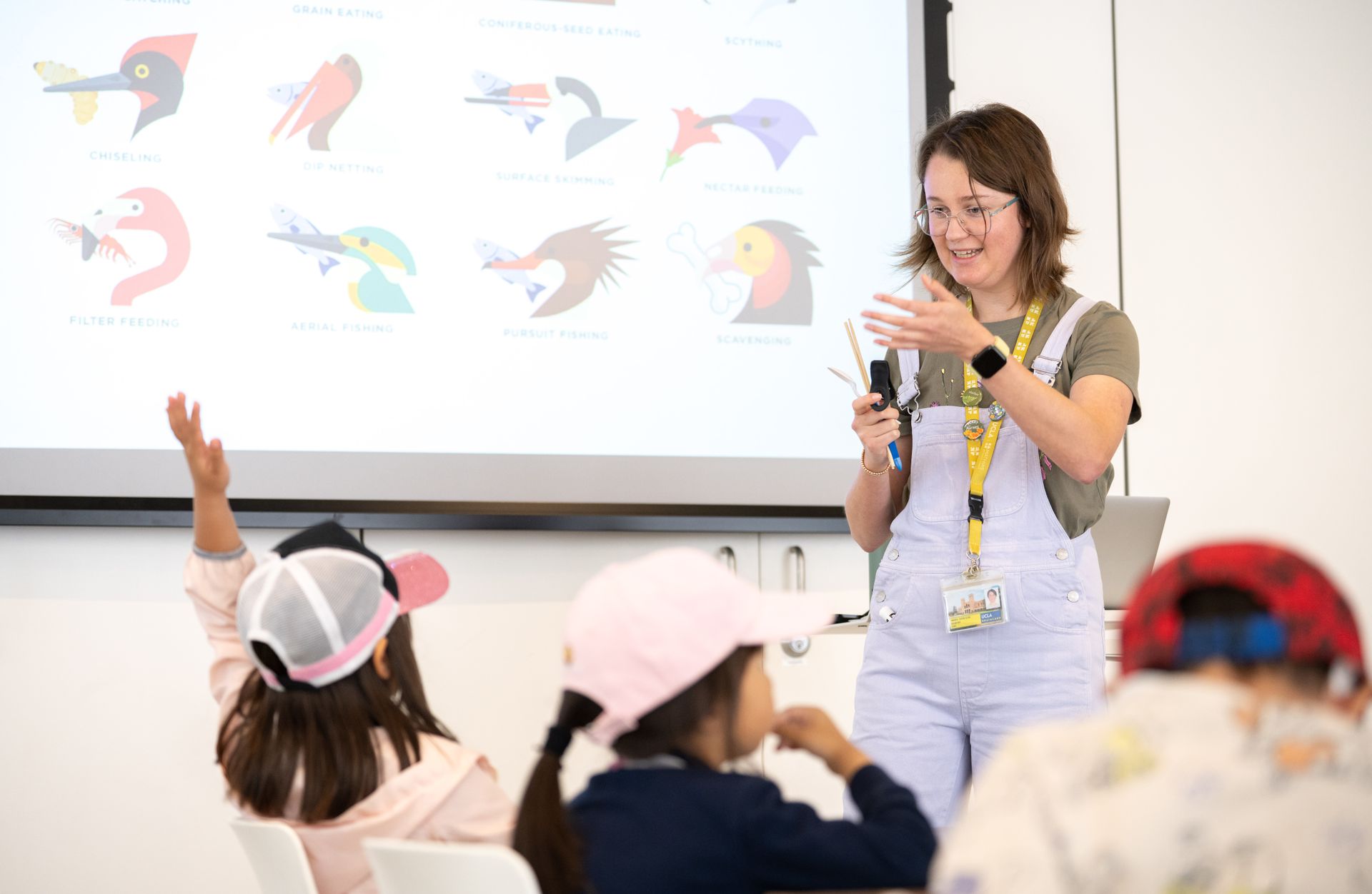 Amanda Bueno-Kling gestures as students raise their hands in front of a screen displaying illustrated bird feeding methods.