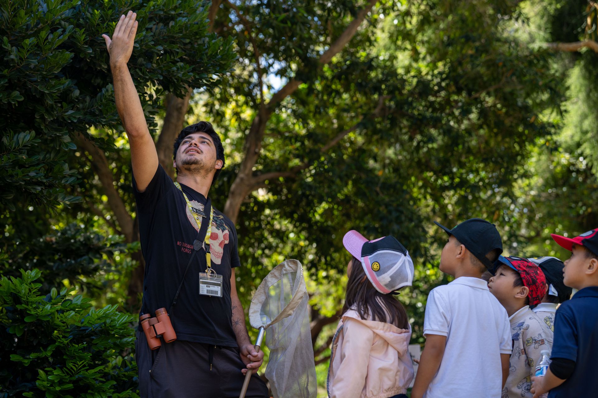 Boaz Solorio leads a group of children on an outdoor tour, pointing up into the trees while holding a butterfly net as the students look on.