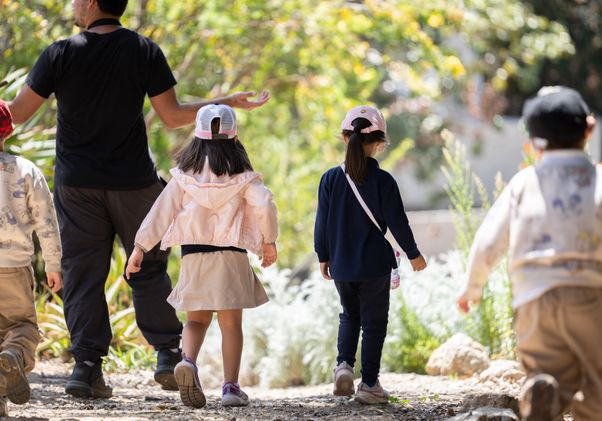 An adult leads a group of children along a sunlit garden path lined with plants and trees during a field trip to the UCLA Mildred E. Mathias Botanical Garden..