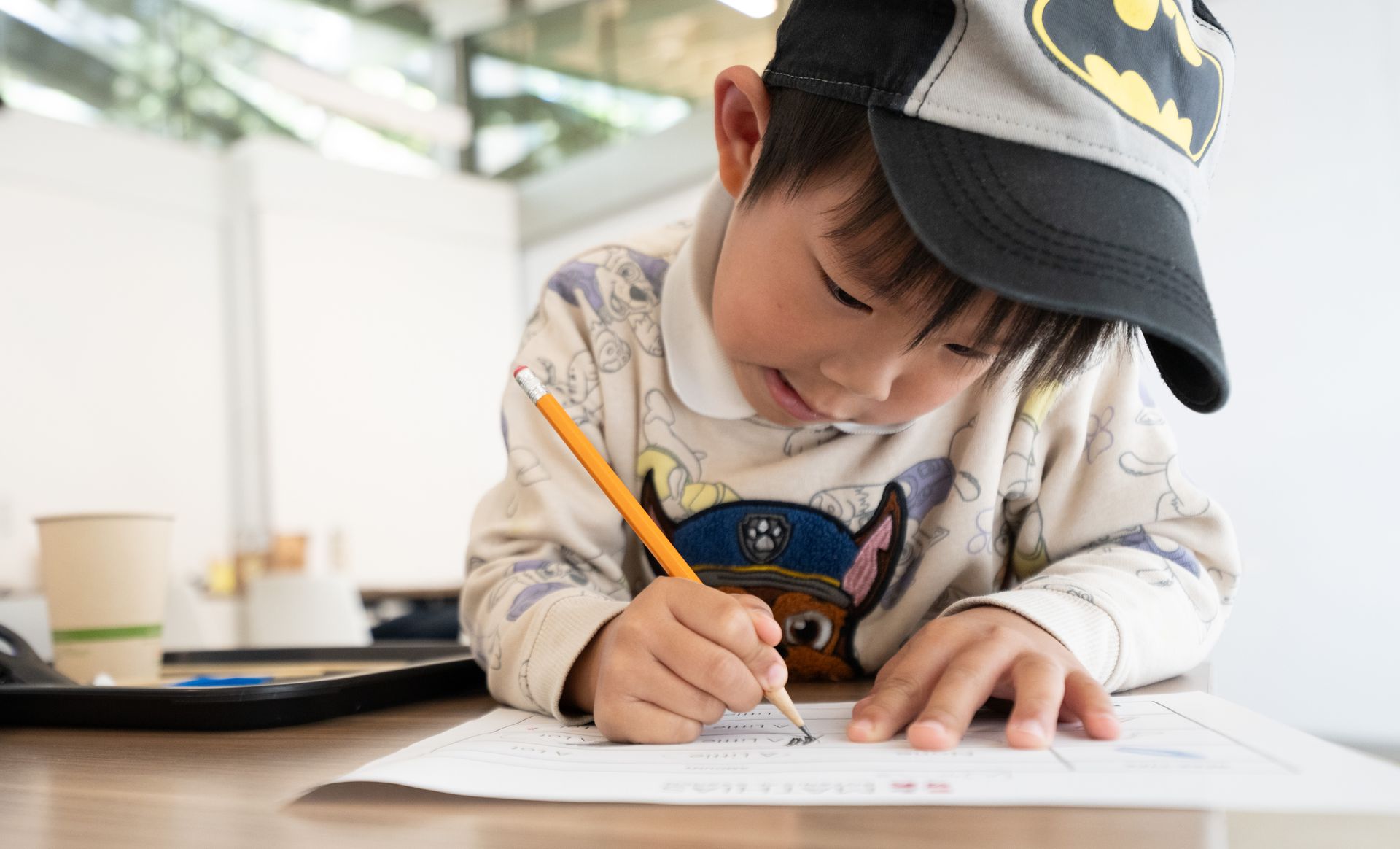 A young child wearing a cap concentrates on writing with a pencil on a worksheet at a classroom desk.