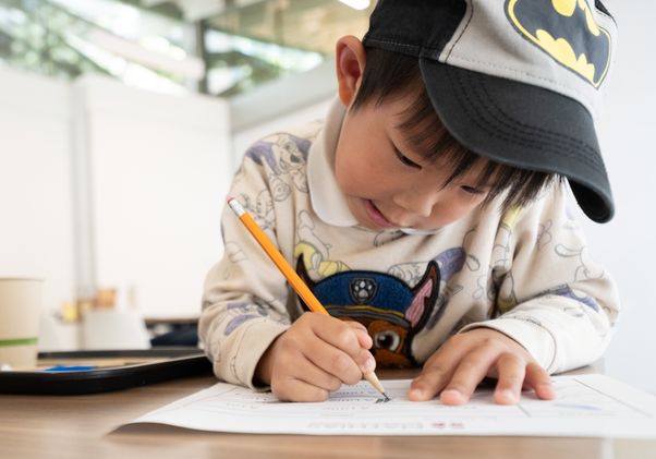 A young child wearing a cap concentrates on writing with a pencil on a worksheet at a classroom desk.