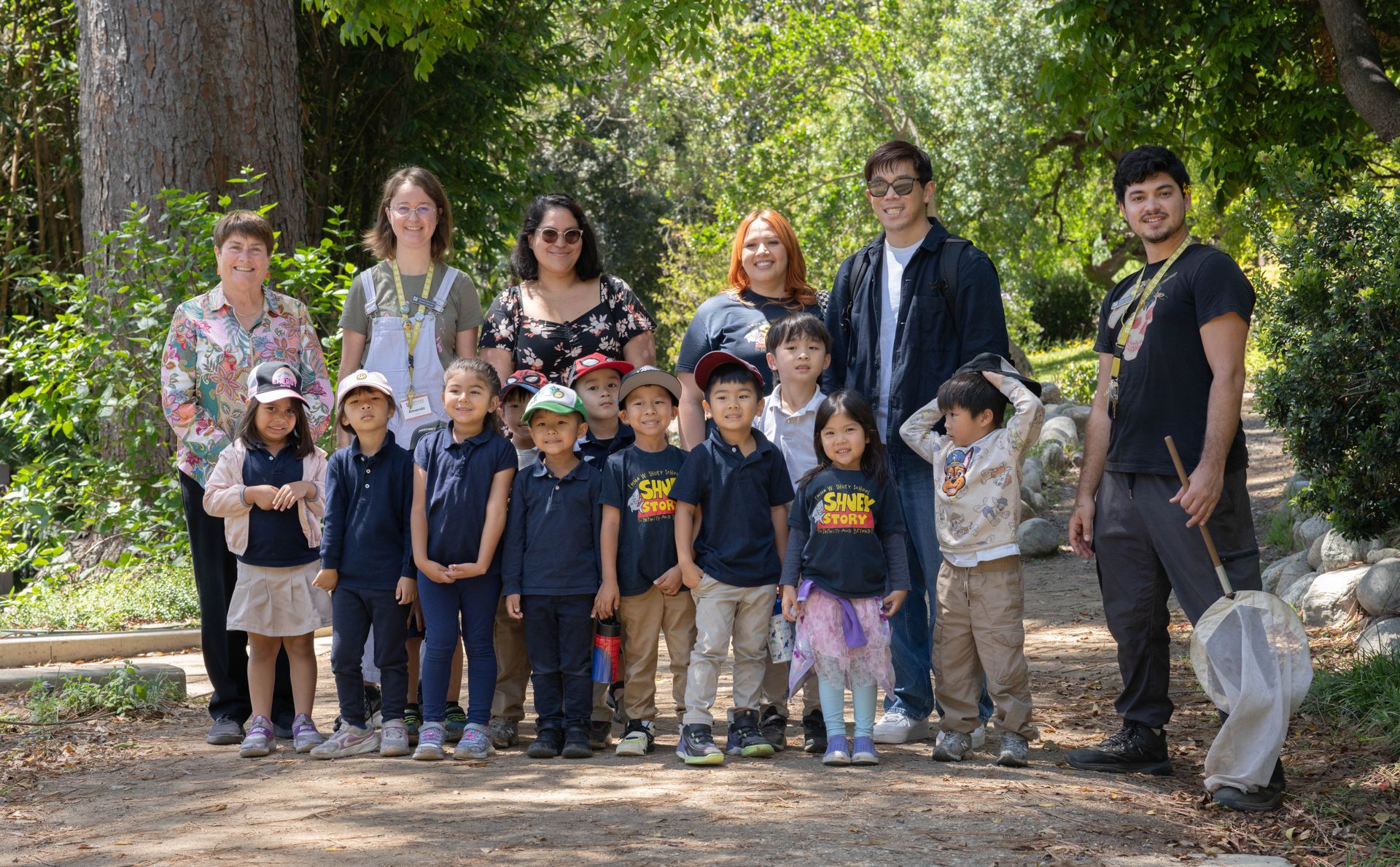 A group of young students and adult guides pose together on a shaded garden path surrounded by trees and greenery.