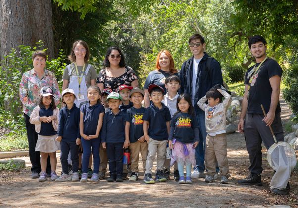 A group of young students and adult guides pose together on a shaded garden path surrounded by trees and greenery.