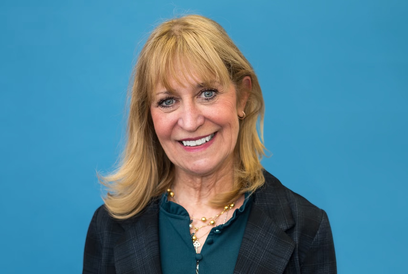 Head-and-shoulders portrait of nursing professor Barbara Bates-Jensen, who wears a dark plaid coat and dark green blouse, against a light blue background.