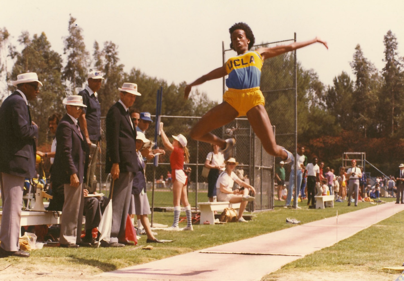 Jackie Joyner-Kersee, in UCLA track-and-field uniform, in mid-air while competing in the long jump; athletics officials look on,