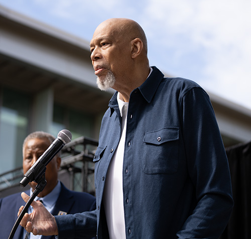 Midrange shot of Kareem Abdul-Jabbar in dark blue shirt standing in front of a microphone
