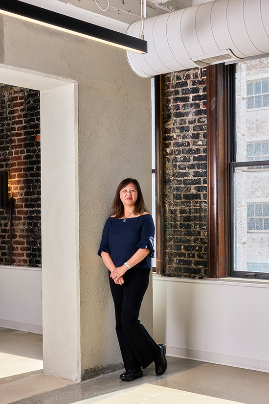 Full shot of Taffany Lim in dark blue top and black pants, standing indoors next to a window