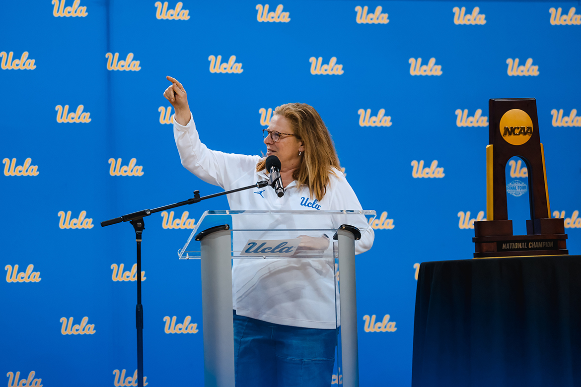 UCLA women's basketball head coach Cori Close stands in front of a UCLA step-and-repeat, gesturing with her right arm, with the NCAA championship trophy on a table to her right.