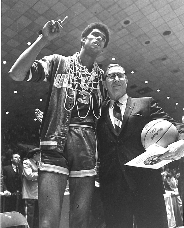 Black-and-white photo of Kareem Abdul-Jabbar standing next to John Wooden and holding up a finger in a No. 1 sign