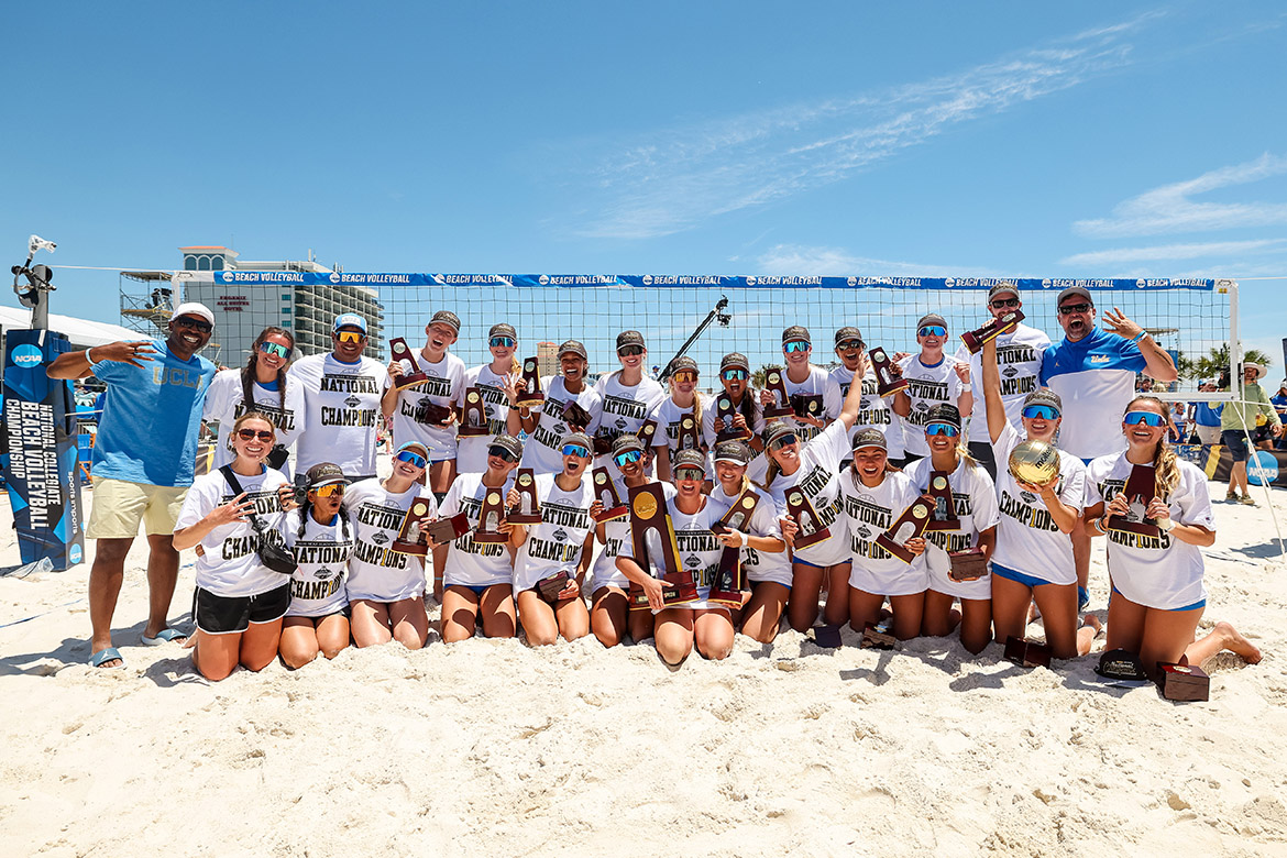 The UCLA women's beach volleyball team and coaching staff pose for a group photo on the sand in front of a beach volleyball net. Players wear white "National Champions" t-shirts and hold individual NCAA trophies and awards.