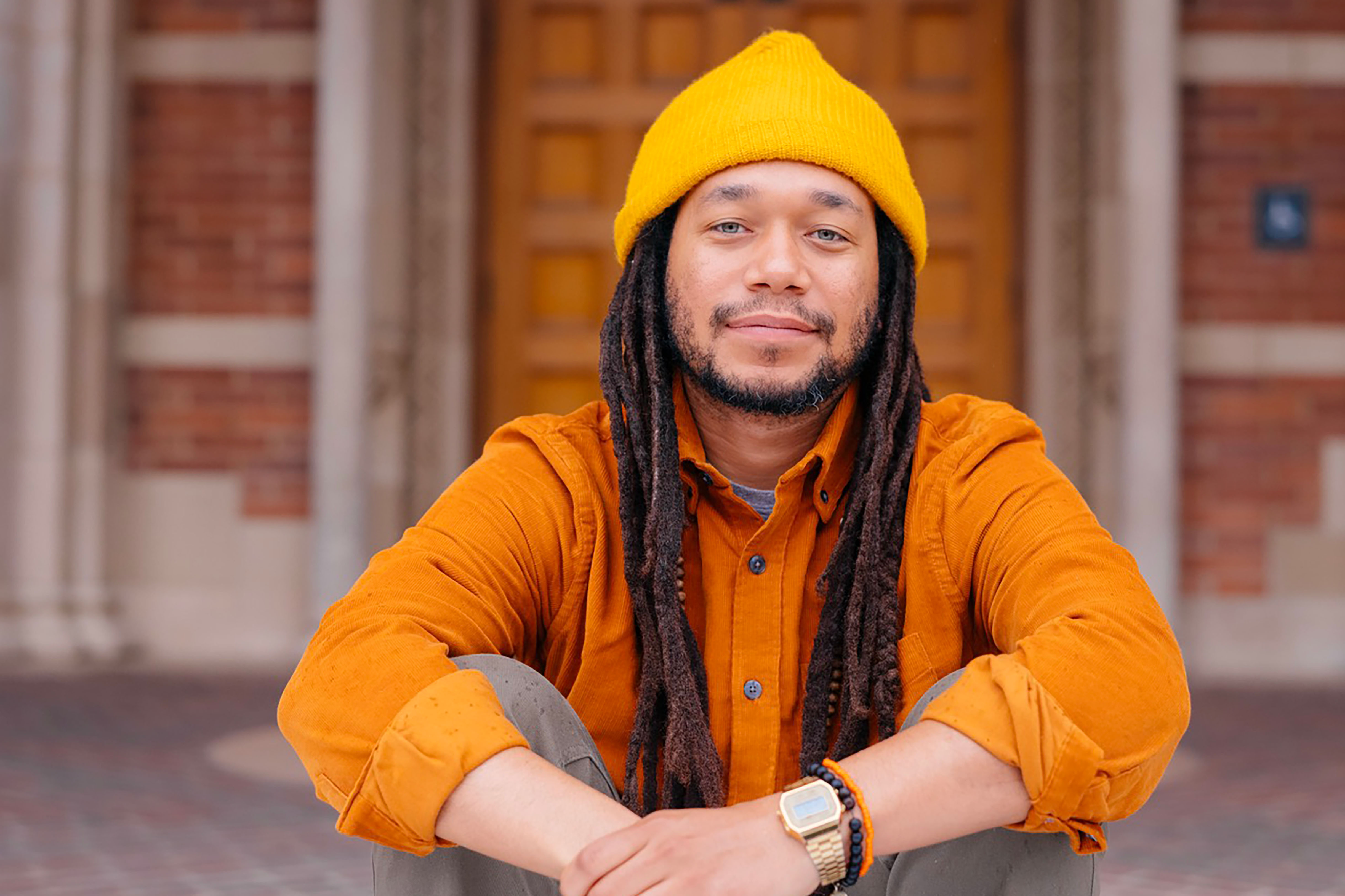 Justin Dunnavant sits cross-legged, wearing a yellow beanie and orange button-down shirt.