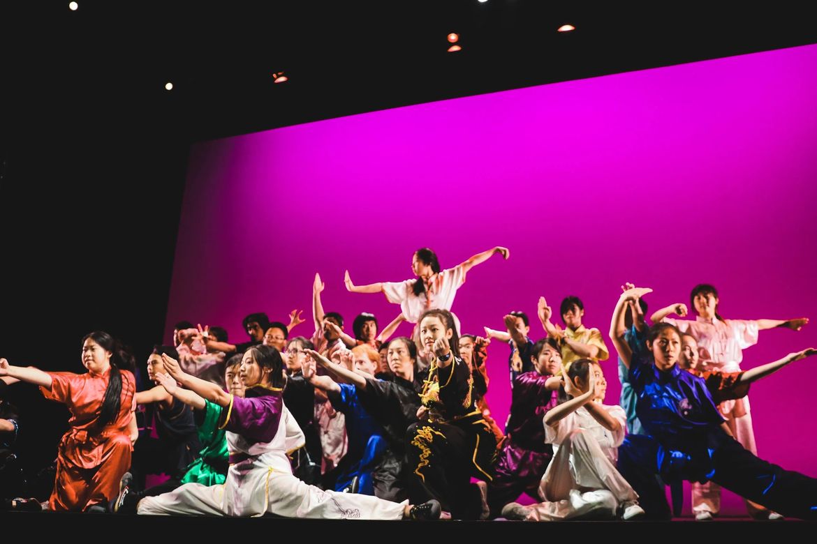 A mass group of Chinese American dancers strike poses on the stage in front of a magenta back wall