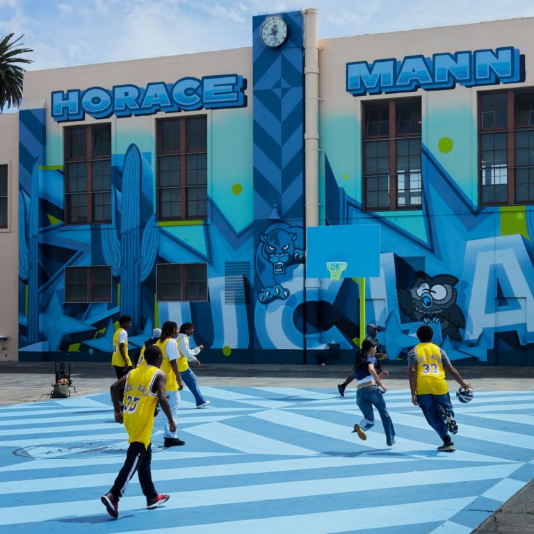 Students play basketball on a blue outdoor court in front of a brightly painted school building reading “Horace Mann.” Several wear yellow jerseys, and a clock is visible above the mural on the building’s facade.