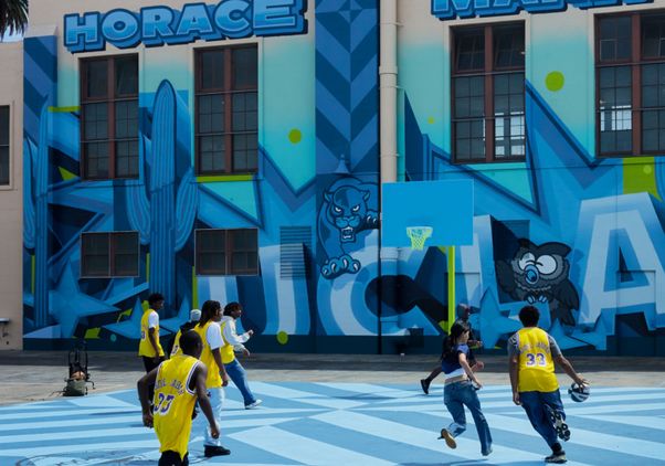 Students play basketball on a blue outdoor court in front of a brightly painted school building reading “Horace Mann.” Several wear yellow jerseys, and a clock is visible above the mural on the building’s facade.
