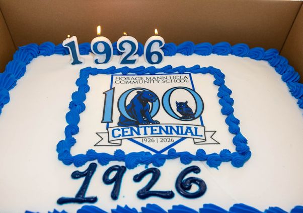 A rectangular cake with blue frosting trim displays “Horace Mann UCLA Community School 100 Centennial 1926–2026.” Four lit candles form “1926” on top, and “1926” is written again in icing below the central centennial logo.