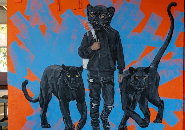Mural by Matthew Grabelsky shows a person with a black panther head, wearing a hoodie and backpack, walking forward between two black panthers. The figures stand against a bright orange wall with blue abstract shapes.