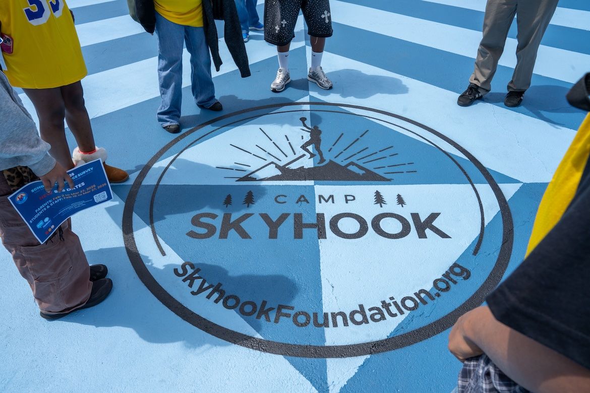 People stand in a circle on a blue court around a large “Camp Skyhook” logo featuring a basketball player silhouette. The text “SkyhookFoundation.org” is visible within the circle.