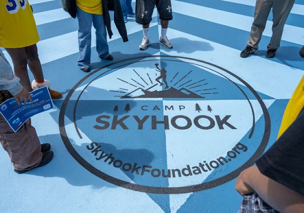 People stand in a circle on a blue court around a large “Camp Skyhook” logo featuring a basketball player silhouette. The text “SkyhookFoundation.org” is visible within the circle.