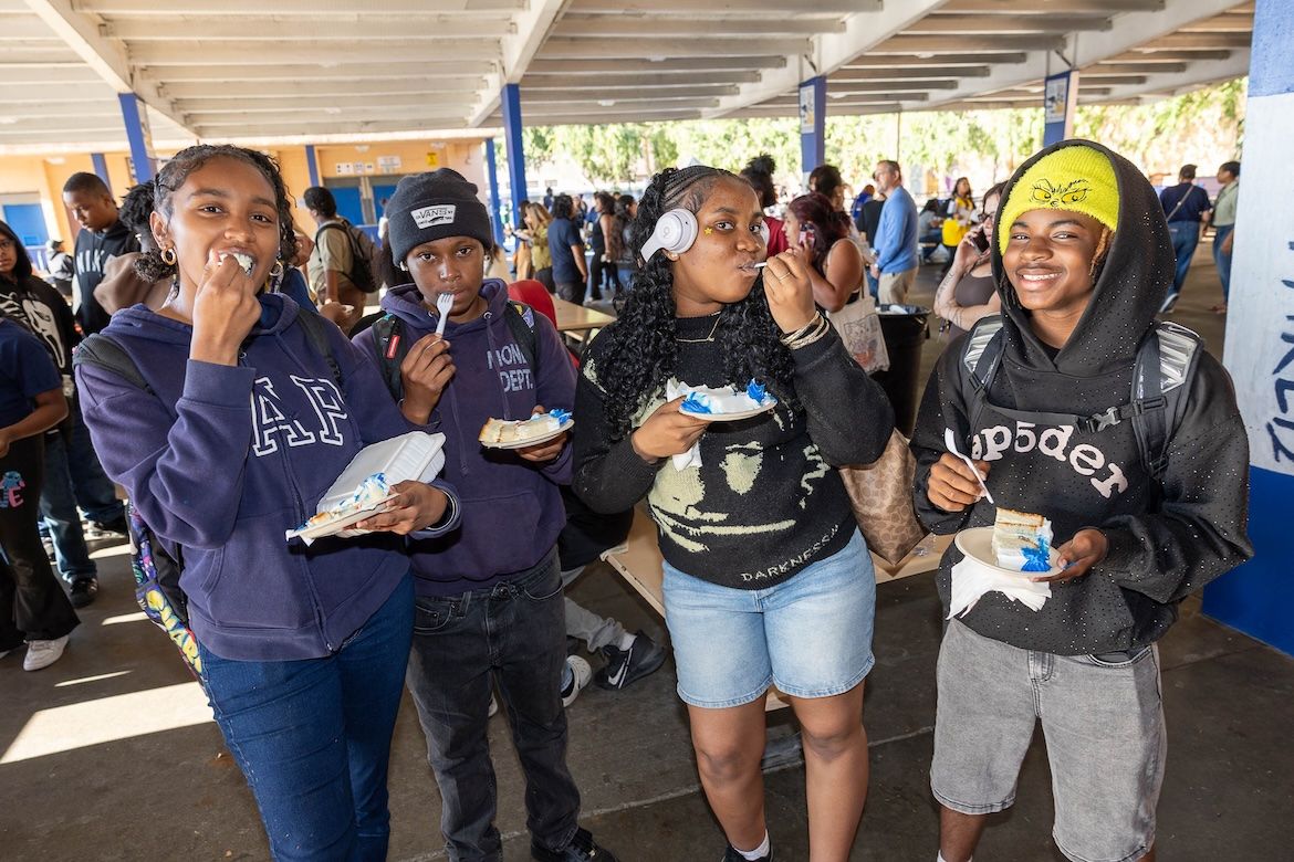 Four Mann school students stand in a covered school courtyard, eating slices of blue-and-white frosted cake from paper plates, smiling and wearing casual clothing and backpacks, with a busy event in the background