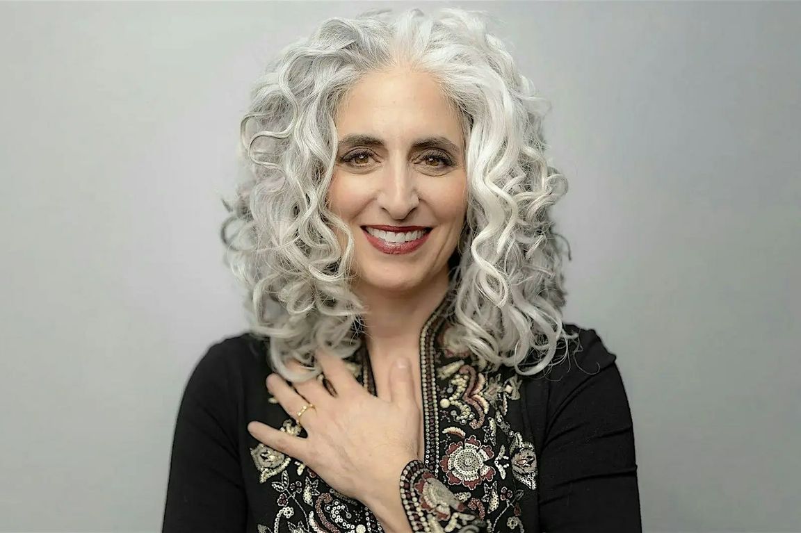 Cantor Elizabeth Shammash smiles in a studio portrait, wearing a patterned garment and resting one hand across her chest.